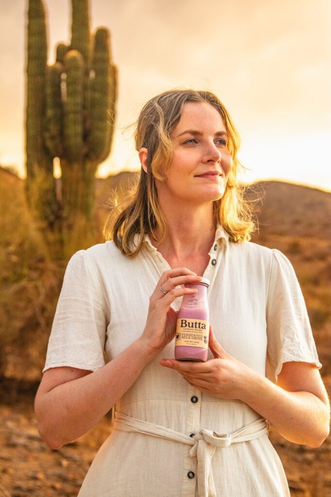 Woman holding a Butta “Crimson Sunset” bottle in the desert at sunset, warm golden light, Arizona landscape